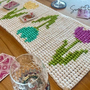 Könnte beinhalten: Weiß gehäkelter Tischläufer mit einem Tulpenmuster in Blau, Gelb, Türkis und Lila. Grüne Stiele und Blätter akzentuieren die Blumen. Spielkarten und ein Glas liegen auf dem Holztisch. Der Text "Sweet Potato 3" befindet sich oben rechts.