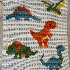 May include: A cream-colored crocheted blanket featuring five colorful dinosaur designs. The dinosaurs are in shades of blue, orange, green, and yellow. The blanket is made of a soft, textured yarn.
