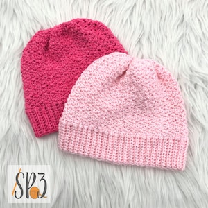 May include: Two crocheted hats, one pink and one hot pink, on a white fuzzy surface.