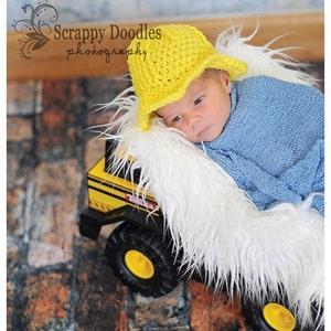 May include: A newborn baby wearing a yellow crocheted hat with a brim, lying on a white fluffy surface. The baby is wearing a blue swaddle and is resting on a yellow toy truck.