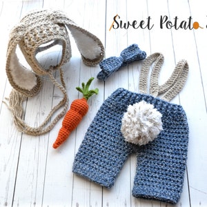 May include: A crocheted bunny costume for a baby, featuring a brown bunny hat with long ears, a blue and white crocheted jumpsuit with suspenders, a white pom-pom tail, and a blue bow. The costume is laid out on a white wooden surface with a crocheted carrot prop.