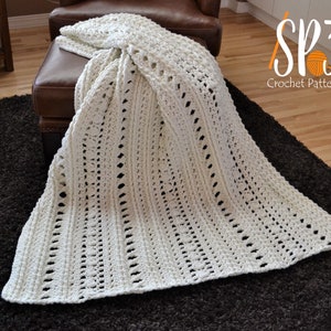 Peut inclure: Une couverture blanche en crochet avec un motif ajouré, drapé sur un ottoman en cuir marron. La couverture est réalisée avec un point délicat et a une texture douce et aérée.