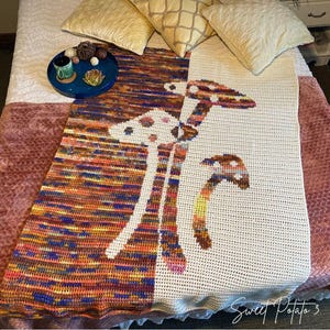 May include: A crochet blanket with a mushroom design. The blanket has a white background with colourful mushroom illustrations. The other half of the blanket has a colourful striped pattern in shades of blue, orange, and purple.