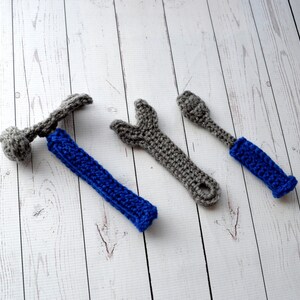 May include: Three crocheted tools, a hammer, wrench, and screwdriver, are displayed on a white wooden surface. The hammer and wrench are gray, and the screwdriver is blue.