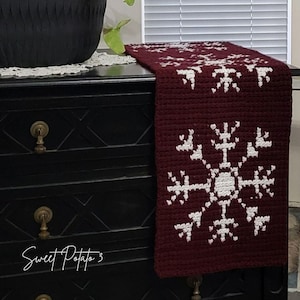 Könnte beinhalten: Burgunderfarbener gehäkelter Tischläufer mit weißem Schneeflockenmuster. Der Läufer liegt auf einer schwarzen Kommode mit goldenen Griffen. Der Text "Sweet Potato 3" ist in Schreibschrift auf die Kommode geschrieben.