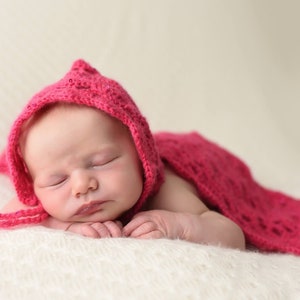 Könnte beinhalten: Ein schlafendes Neugeborenes, das eine rosa gestrickte Kapuze trägt. Das Baby liegt auf einer weißen Decke.