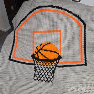 Könnte beinhalten: Eine graue Häkeldecke mit einem Basketballkorb-Design. Der Reifen ist schwarz und orange umrandet, mit einem orangefarbenen Basketball und einem schwarzen Netz. Das Wort "HOME" ist im Hintergrund sichtbar.