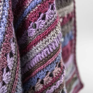 Puede incluir: Primer plano de una manta de ganchillo con un patrón de rayas multicolores en tonos de morado, rosa, azul y marrón.