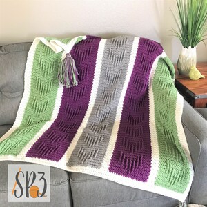 Peut inclure: Une couverture en crochet rayée dans les tons de vert, violet, gris et blanc. La couverture est posée sur un canapé gris et a un pompon attaché à un coin.