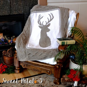 Peut inclure: Une couverture en crochet avec un fond blanc et une silhouette de cerf marron au centre. La couverture est drapé sur un fauteuil en bois avec un livre sur l'accoudoir.