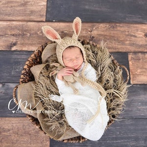 May include: A newborn baby wearing a crocheted bunny hat and a white swaddle is sleeping in a wicker basket lined with a brown faux fur blanket. The baby is holding a crocheted bunny toy.