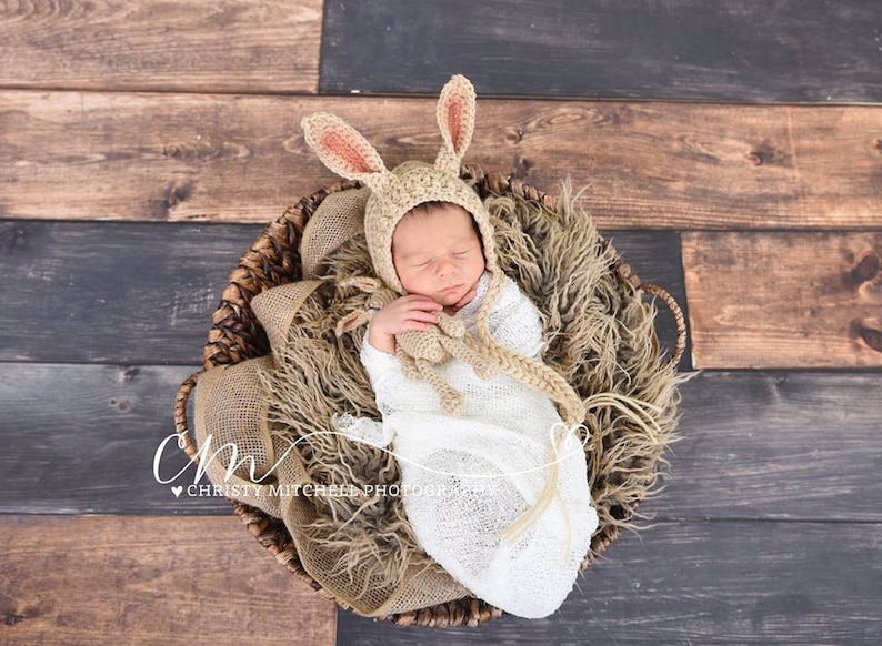 May include: A newborn baby wearing a crocheted bunny hat and a white swaddle is sleeping in a wicker basket lined with a brown faux fur blanket. The baby is holding a crocheted bunny toy.