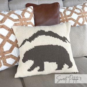 Può includere: Un cuscino bianco all'uncinetto con un design a silhouette di orso marrone. Il cuscino è su un divano con altri cuscini a motivi geometrici.
