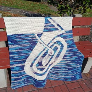 May include: A rectangular crochet blanket featuring a large, white, stylized tuba graphic centered against a variegated background of blue, teal, and purple yarn. The top section of the blanket is solid white, and the text Sweet Potato 3 is visible.