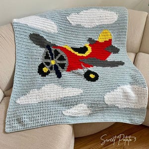 Puede incluir: Manta de bebé tejida a ganchillo con un avión rojo con detalles amarillos y negros, alas grises y una hélice. El fondo es azul claro con nubes blancas. El texto "Sweet Potato 3" está en la esquina inferior derecha.