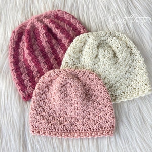 May include: Three crocheted hats in pink, cream, and a striped pink and white pattern. The hats are arranged on a white fluffy surface.