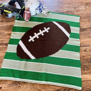 Könnte beinhalten: Eine handgefertigte Häkeldecke mit einem Football-Design. Die Decke zeigt einen braunen Football mit weißen Streifen auf einem grün-weiß gestreiften Hintergrund. Ein Football-Helm und -Polster befinden sich im Hintergrund.