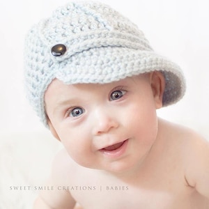 May include: A light blue crocheted baby hat with a small brim and a black button. The hat is worn by a baby with blue eyes. The text "SWEET SMILE CREATIONS | BABIES" is at the bottom of the image.
