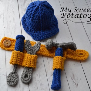 Pode incluir: Um chapéu de trabalhador da construção civil de crochê azul com um cinto de ferramentas de crochê amarelo com uma chave de fenda, uma chave e um martelo.