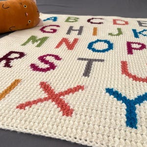 Könnte beinhalten: Eine cremefarbene, gehäkelte Decke mit dem Alphabet in verschiedenen Farben. Die Buchstaben sind in einem kräftigen Blockstil gestickt. Ein plüschiges, orangefarbenes, katzenförmiges Kissen liegt auf der Decke.