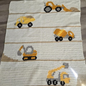May include: A cream-colored crochet blanket featuring yellow construction vehicles: a dump truck, excavator, cement mixer, and a front-end loader. The blanket has horizontal tan stripes and the text "Sweet Potato 3" in the upper left corner.