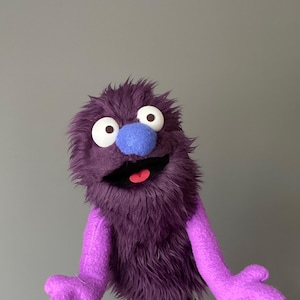 May include: A purple puppet with a shaggy, furry body and large white eyes. It has a blue nose and a wide, open mouth revealing a red tongue. The puppet's arms are a lighter shade of purple.