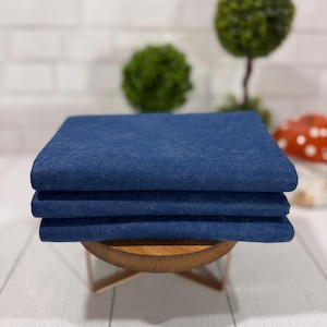 May include: Three stacks of indigo blue fabric, neatly folded and stacked on a small, round wooden stand. The fabric has a textured appearance, and the color is a deep, saturated blue. The background includes decorative greenery and a white brick wall.