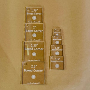 May include: A set of nine clear acrylic boxed corner templates in various sizes, ranging from 0.75 inches to 2.5 inches. Each template features the text "Boxed Corner" and the size in inches. The templates are stacked on a brown surface.
