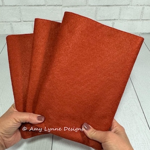 May include: Three sheets of orange felt fabric folded together. The felt is a deep, rich orange color.  © Amy Lynne Designs