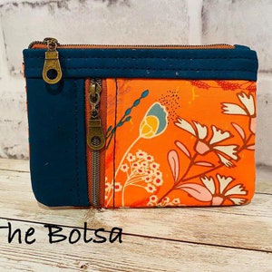 May include: A small orange zippered pouch with a floral print and a dark blue fabric panel. The pouch has two zippers and a brass zipper pull. The text "The Bolsa" is written on the wooden surface below the pouch.