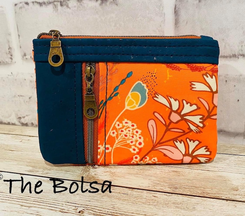 May include: A small orange zippered pouch with a floral print and a dark blue fabric panel. The pouch has two zippers and a brass zipper pull. The text "The Bolsa" is written on the wooden surface below the pouch.