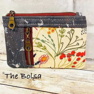 May include: A small zippered pouch with a floral print on one side and a cork fabric on the other side. The pouch has two zippers and a brown metal zipper pull. The pouch is sitting on a wooden surface. The text "The Bolsa" is visible in the bottom left corner of the image.
