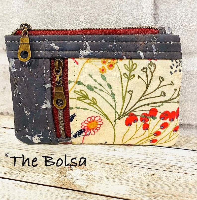 May include: A small zippered pouch with a floral print on one side and a cork fabric on the other side. The pouch has two zippers and a brown metal zipper pull. The pouch is sitting on a wooden surface. The text "The Bolsa" is visible in the bottom left corner of the image.