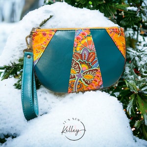 Pode incluir: Uma carteira de mão de formato de meia lua azul turquesa com um painel de tecido estampado com flores. A carteira tem uma alça azul turquesa e um zíper dourado. A carteira está sobre um leito de neve.