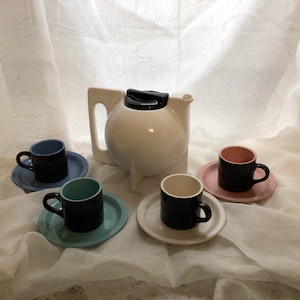 Può includere: Una teiera in ceramica bianca con un coperchio nero e un corpo rotondo, insieme a quattro tazze e piattini in vari colori: blu, turchese, rosa e bianco.