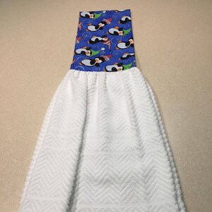 May include: A white kitchen towel with a chevron pattern. The top portion features a blue fabric with a pattern of penguins wearing hats and scarves. The penguins are black, white, and orange.