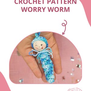 Könnte beinhalten: Ein blauer und weißer gehäkelter Sorgenwurm mit einem Holzkopf und einem lächelnden Gesicht. Der Wurm wird in einer Hand gehalten. Der Text "BABSI.AT" und ein Logo für "babsis" sind im Bild zu sehen.