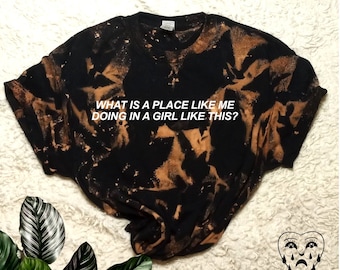 Könnte beinhalten: Schwarzes T-Shirt mit braunen Bleichmittelspritzern und dem Text "WHAT IS A PLACE LIKE ME DOING IN A GIRL LIKE THIS?" in Weiß. Das Shirt liegt auf einer weißen, flauschigen Oberfläche. Der Text "Avery Shane Co." befindet sich in der rechten unteren Ecke des Bildes.