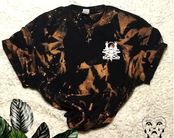 Könnte beinhalten: Schwarz gebleichtes T-Shirt mit einem weißen Grafikdruck eines Dreiecks mit einem Auge darin und zwei Figuren auf jeder Seite. Die Grafik ist mittig auf dem Shirt platziert. Das Shirt hat einen Used-Look.