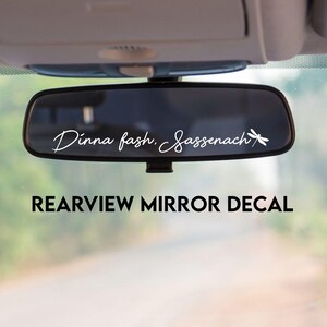 May include: A rearview mirror decal with the text "Dinna fash, Sassenach" in a script font, with a dragonfly graphic. The decal is displayed inside a car, with a blurred outdoor background.