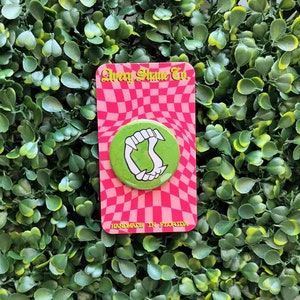 May include: A green button with white fangs on a pink and red checkered background. The button is attached to a pink card with the text "Avery Shane Co." and "HANDMADE IN FLORIDA". The background is a green, leafy surface.
