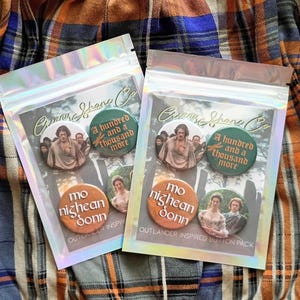 May include: A set of four Outlander inspired button pins. The buttons feature characters from the show, including Jamie Fraser, Claire Fraser, and a quote from the show. The buttons are packaged in a clear plastic bag with a white background and the text "Outlander Inspired Button Pack".