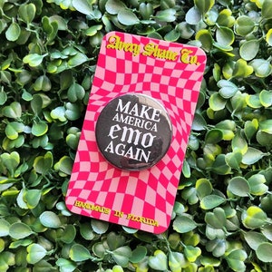 May include: Black button with white text that reads "Make America Emo Again" on a pink and white checkered background. The background also has the text "Avery Shine Co." and "Handmade in Florida".