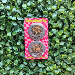 May include: Two round, black and gold door knocker pins with a whimsical, cartoon-like design. The pins are on a pink and white checkered background with the text "Avery Shane Co. Handmade in Florida".