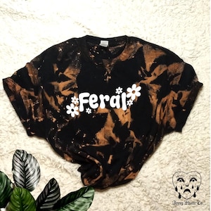 Könnte beinhalten: Schwarzes T-Shirt mit einem Bleich-Tie-Dye-Muster. Das Shirt trägt den Schriftzug "Feral" in Weiß mit weißen Blumen drumherum.