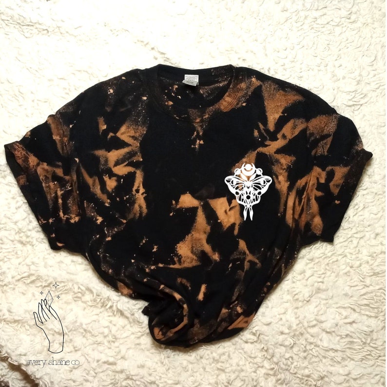 Op de afbeelding: Zwart T-shirt met een bruin gebleekt tie-dye patroon. Een witte afbeelding van een nachtvlinder met een halve maan en sterren is gecentreerd op het shirt.