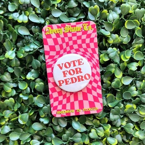 May include: A white button with the text "VOTE FOR PEDRO" in red and orange letters on a pink and white checkered background. The button is labeled "Avery Shane Co. Handmade in Florida."