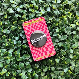 May include: Black and white button with the word "Bleighssed" in a gothic font. The button is on a pink and white checkered background with the text "Away Shine Co. Handmade in Florida".