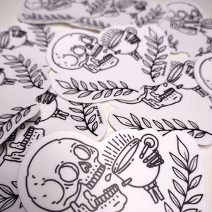 May include: A black and white sticker featuring a skull with a cup of coffee pouring into its mouth. The sticker has a simple line art style and includes a leafy branch design. The text "Deny Sleep Co." is at the bottom of the sticker.