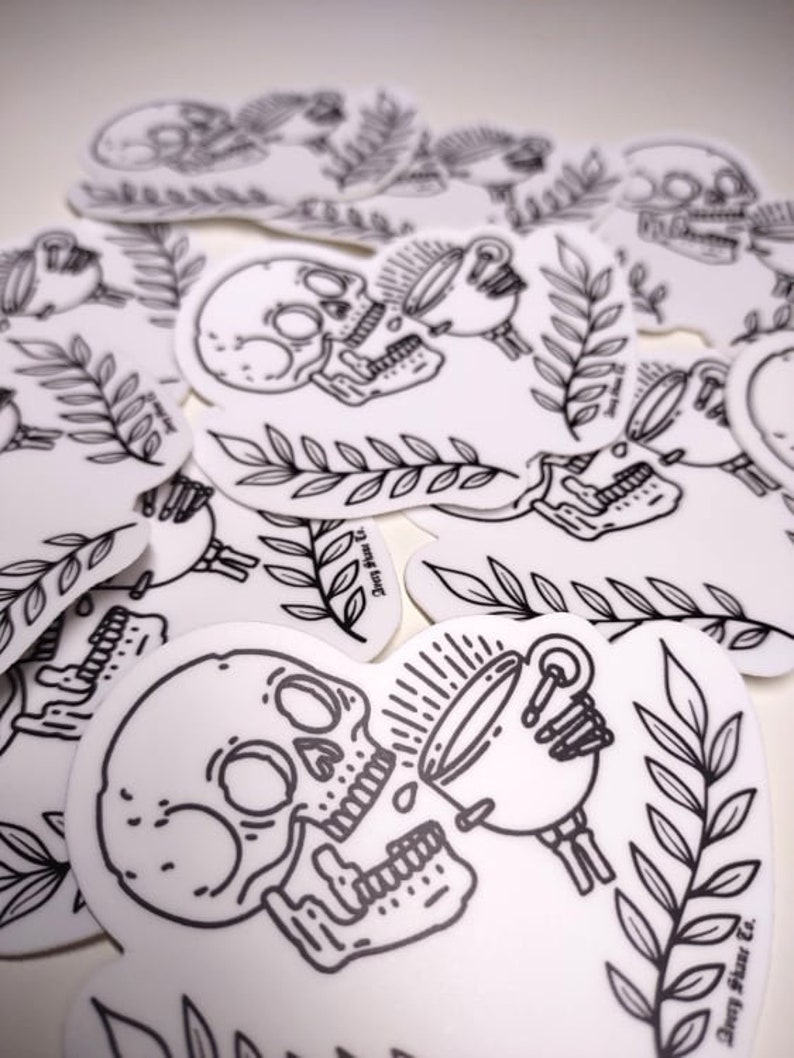 May include: A black and white sticker featuring a skull with a cup of coffee pouring into its mouth. The sticker has a simple line art style and includes a leafy branch design. The text "Deny Sleep Co." is at the bottom of the sticker.